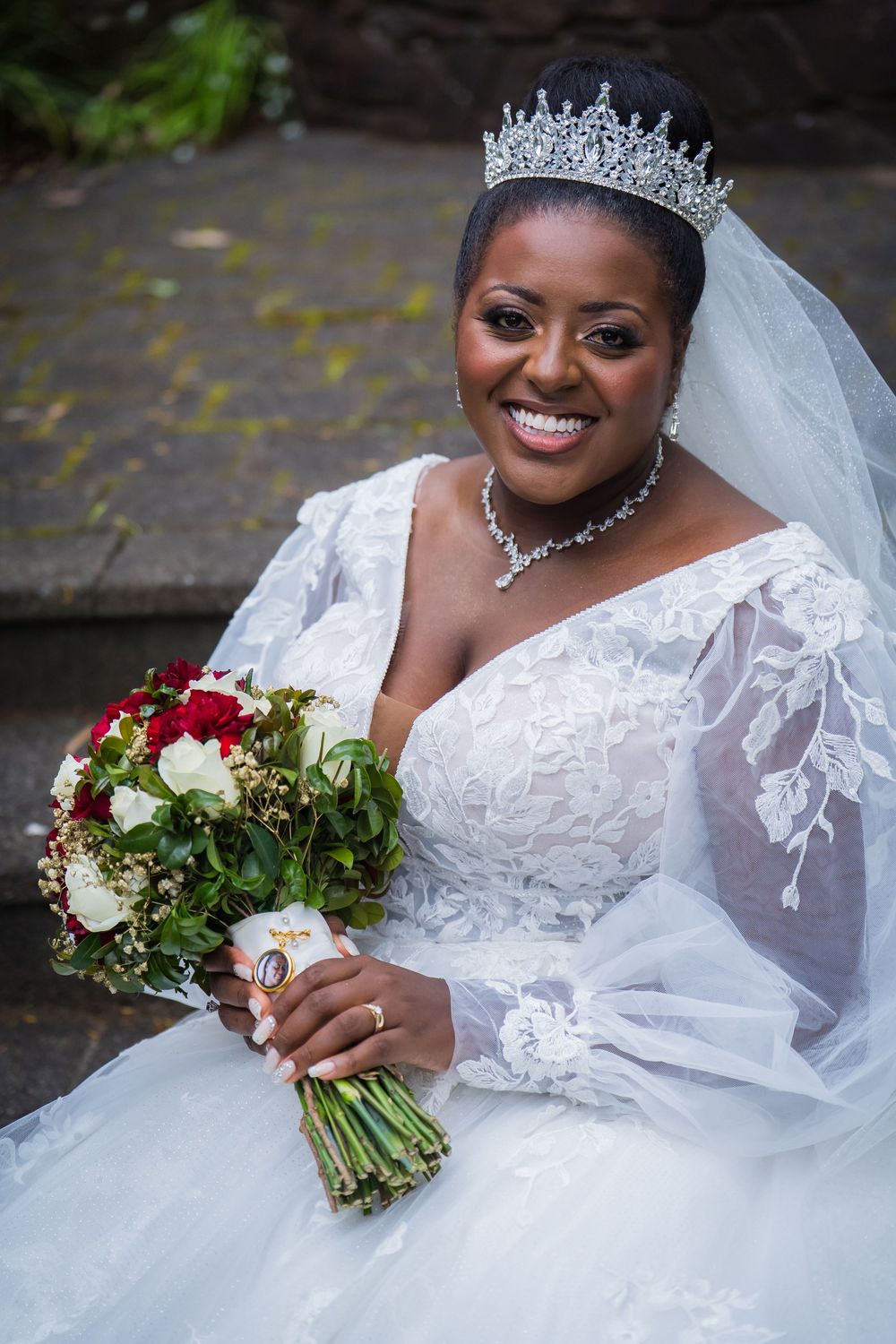 Bride in white lace wedding dress with tiara and veil holds red and white bouquet in outdoor setting.
