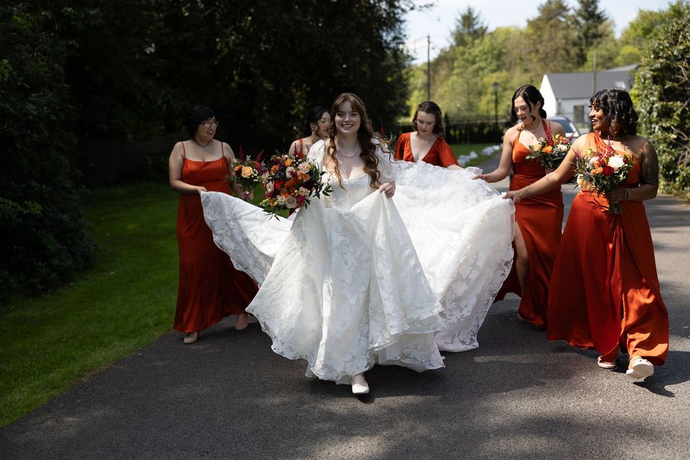 A bride in a flowing white gown walks with bridesmaids in burnt orange dresses on a tree-lined path. Cabra Castle