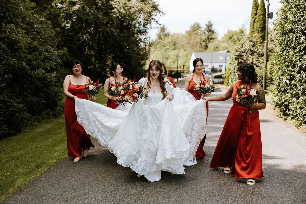 Bridesmaids in burgundy dresses help carry a white wedding gown train along a sunlit garden pathway. Cabra Castle