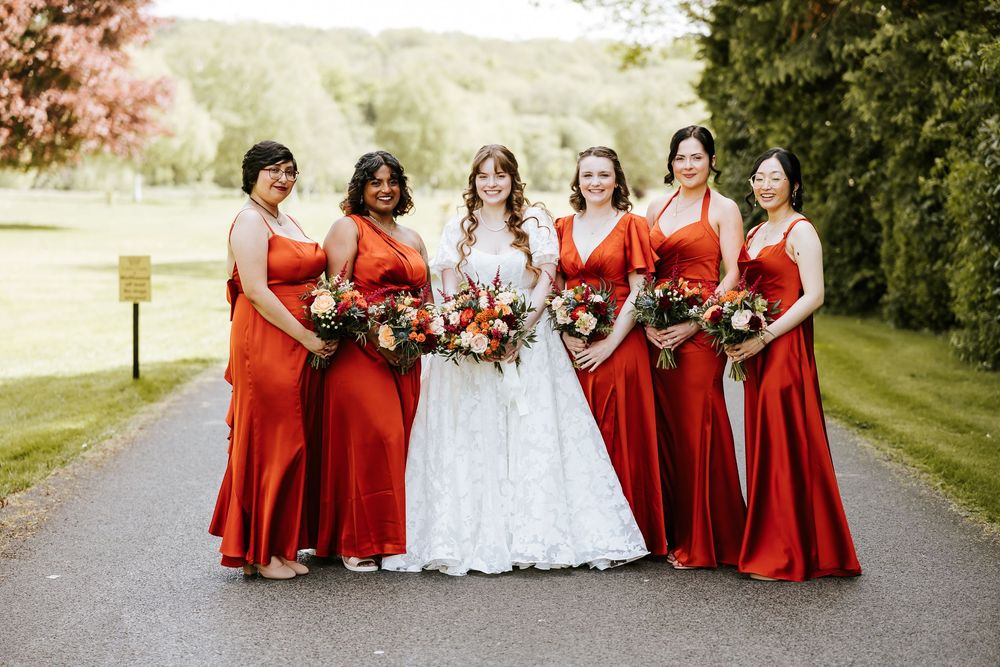 A wedding party wearing vibrant red dresses poses together on a tree-lined path in an outdoor setting. Cabra Castle
