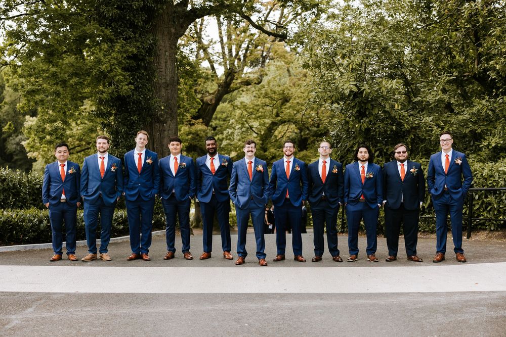 Groomsmen in matching navy blue suits stand in a row outdoors against a lush green garden backdrop. Cabra Castle