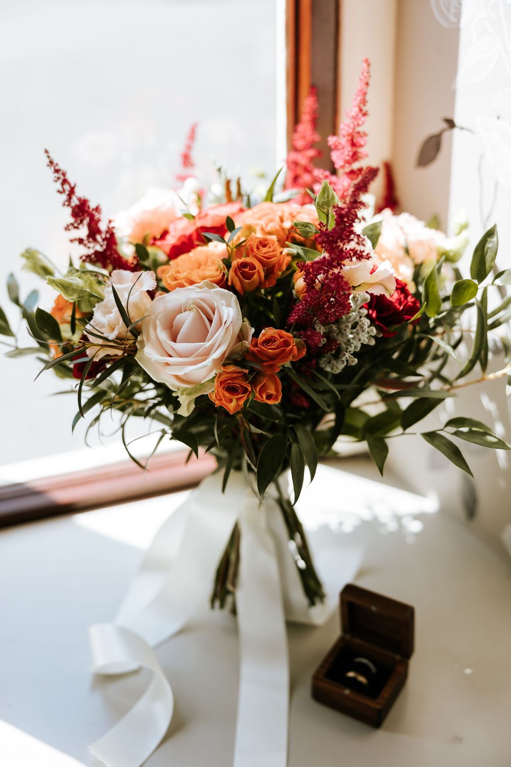 A romantic wedding bouquet with white roses, orange spray roses, red astilbe and greenery sits beside a ring box in sunlight. Cabra Castle