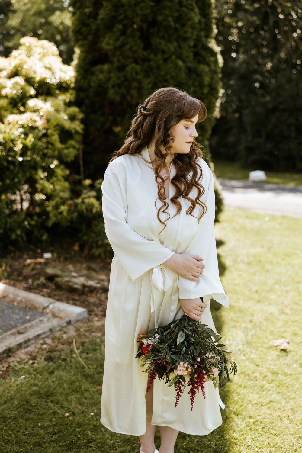 Woman in white robe holding dark floral bouquet poses gracefully in outdoor garden setting. Cabra Castle