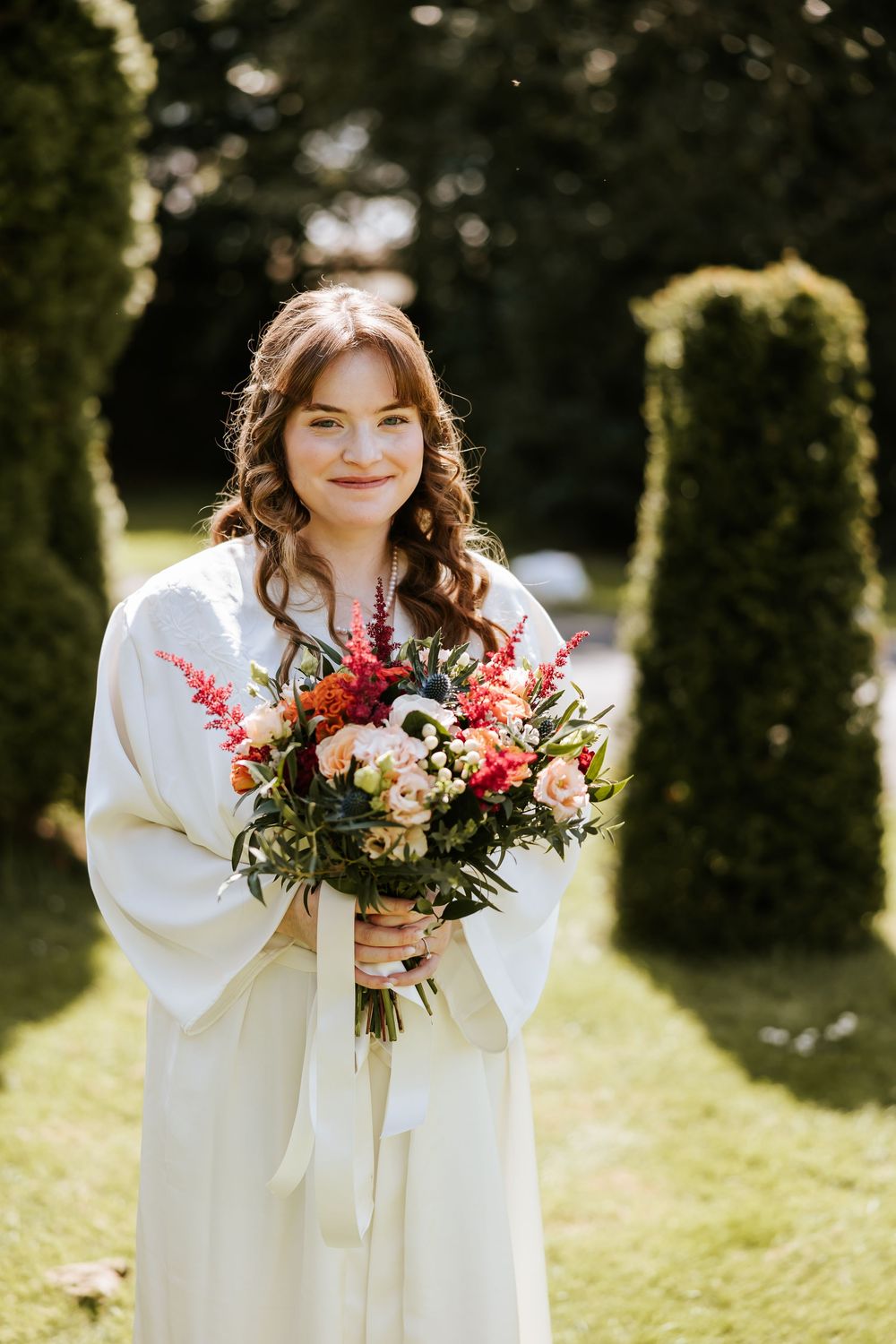 A person in a white dress holds a vibrant bouquet of red and pink flowers in a sunny garden setting. Cabra Castle
