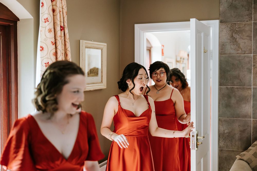 A group of bridesmaids in red dresses share a joyful moment together while getting ready in a sunlit room. Cabra Castle