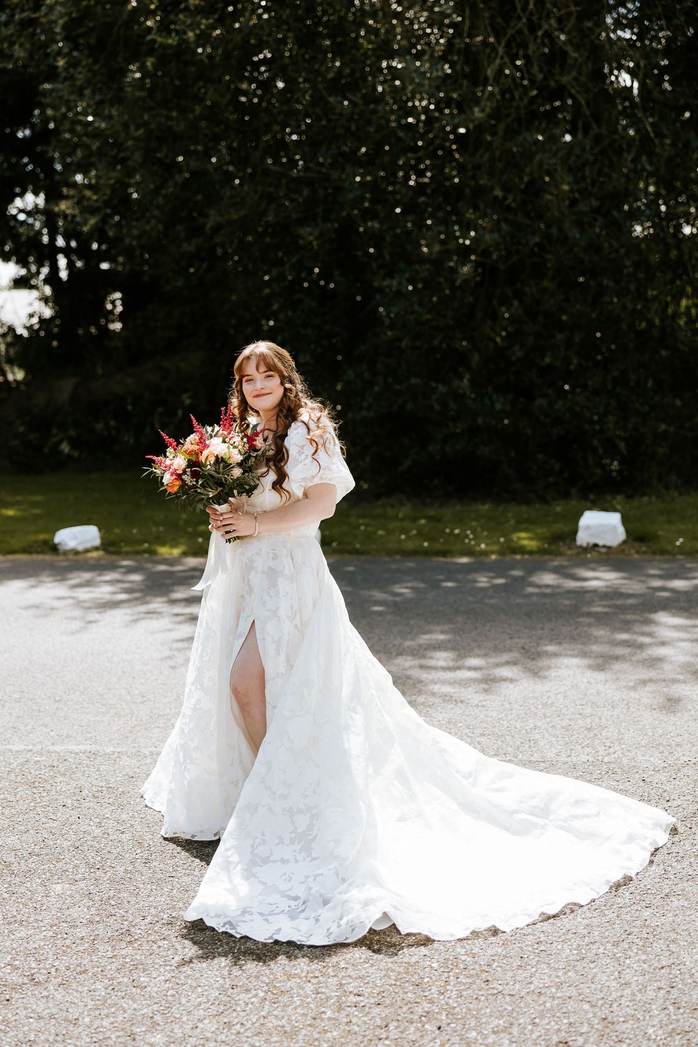 A bride in a flowing white lace gown twirls gracefully on a gravel path surrounded by green foliage. Cabra Castle