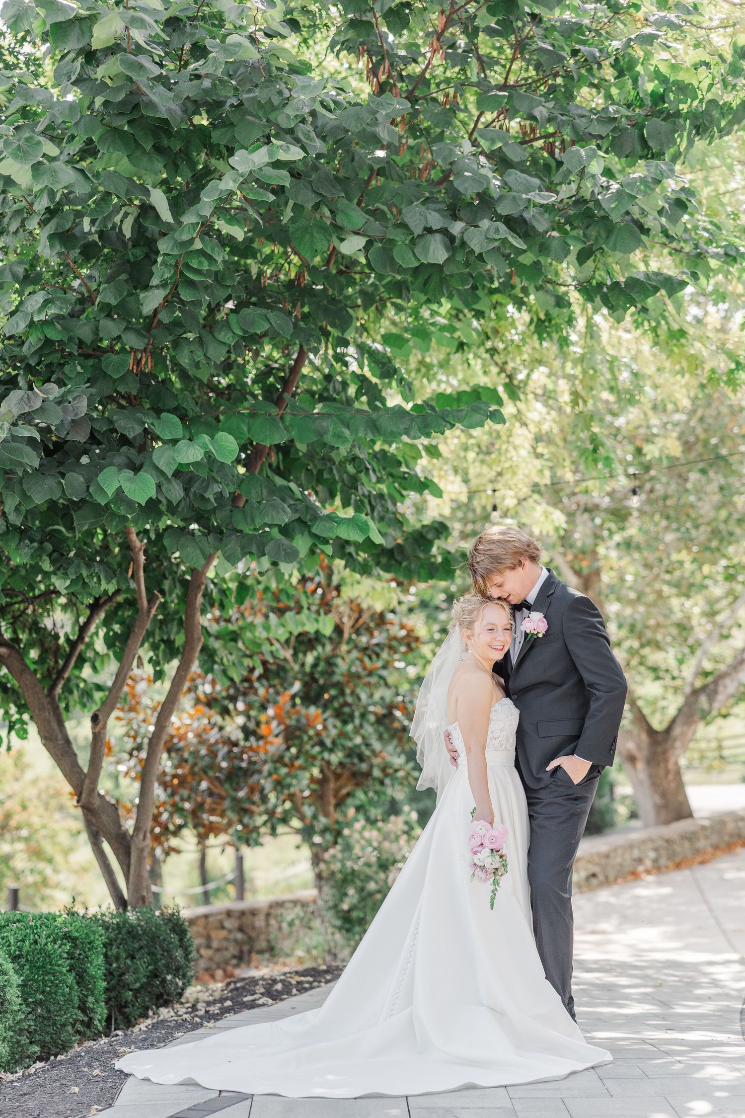A romantic wedding portrait of a couple embracing under a leafy green tree.