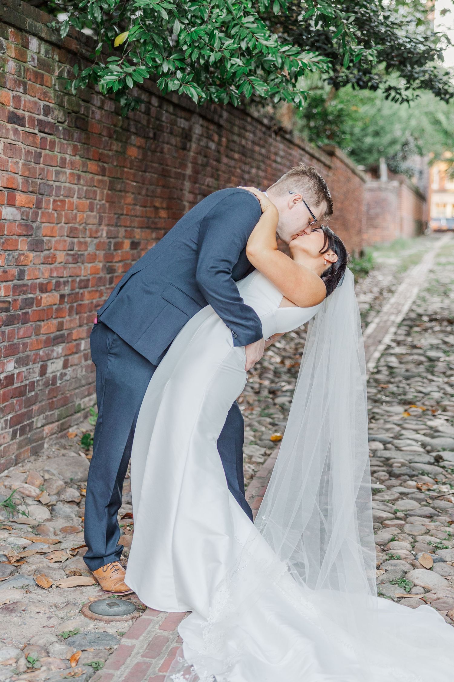 A romantic wedding couple shares a dip kiss on a cobblestone path beside a rustic brick wall.