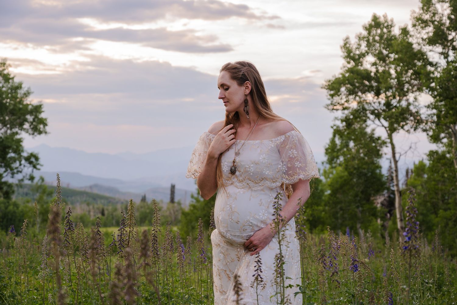 Pregnant woman in white lace dress standing in wildflower field at sunset in mountains.