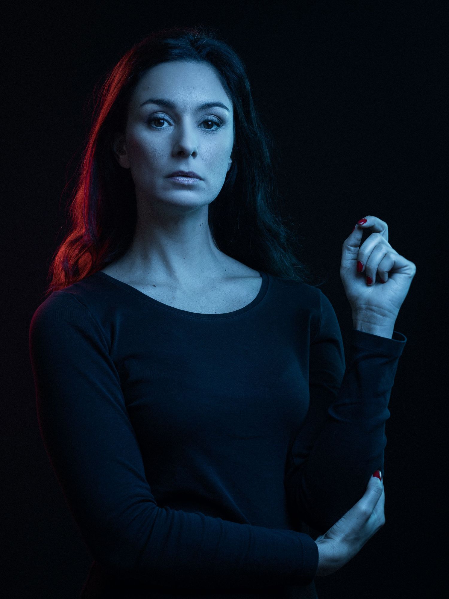 Woman in black long-sleeve top poses dramatically against dark background with red and blue lighting.
