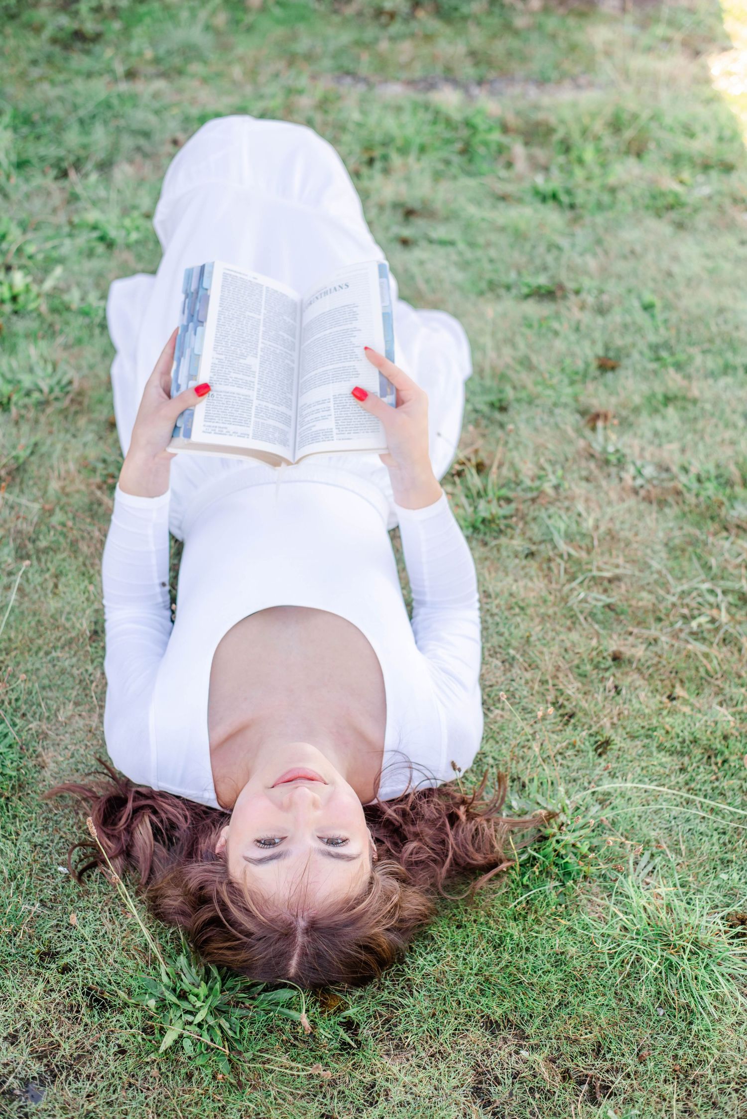 Woman in white dress lying on grass reading an open book outdoors on a sunny day.