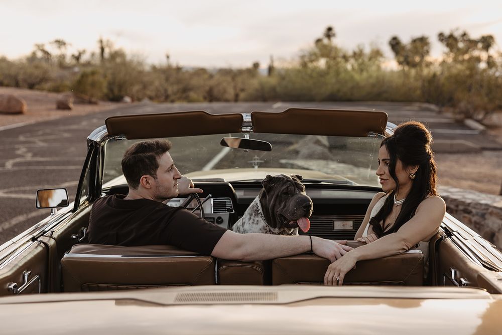 Man and woman with their dog sitting together in a classic convertible car in a desert setting.