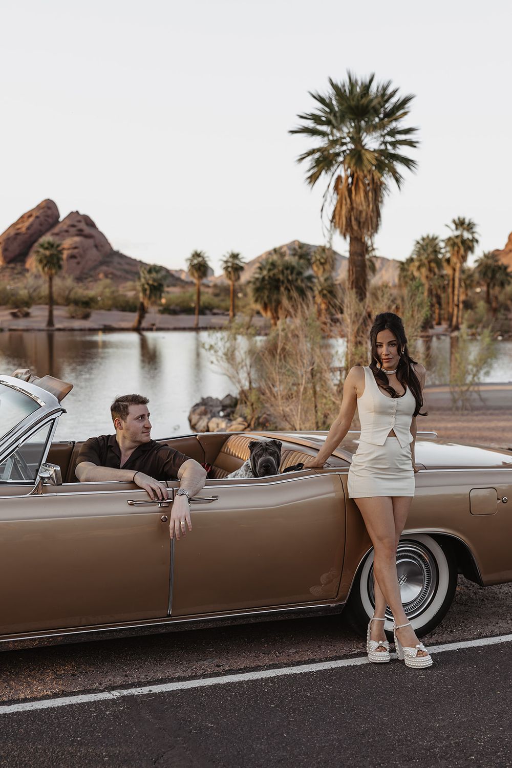 Couple posing with a vintage tan convertible car near a lake with palm trees and rocky hills in the background.