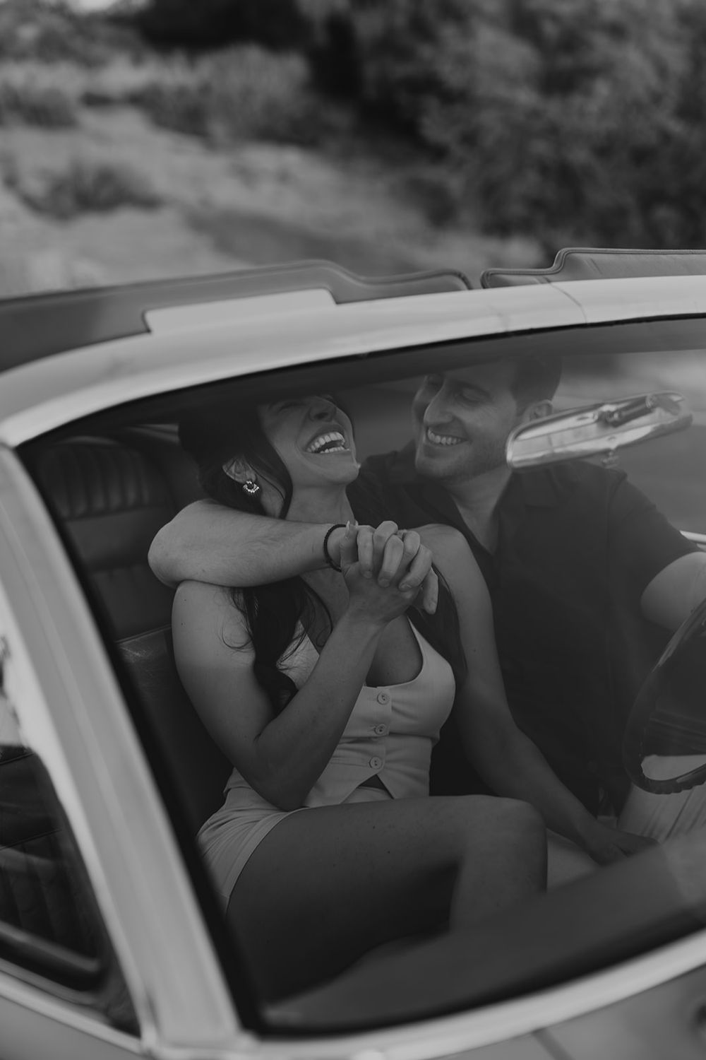 Black and white photo of a happy couple holding hands and laughing together inside a convertible car.