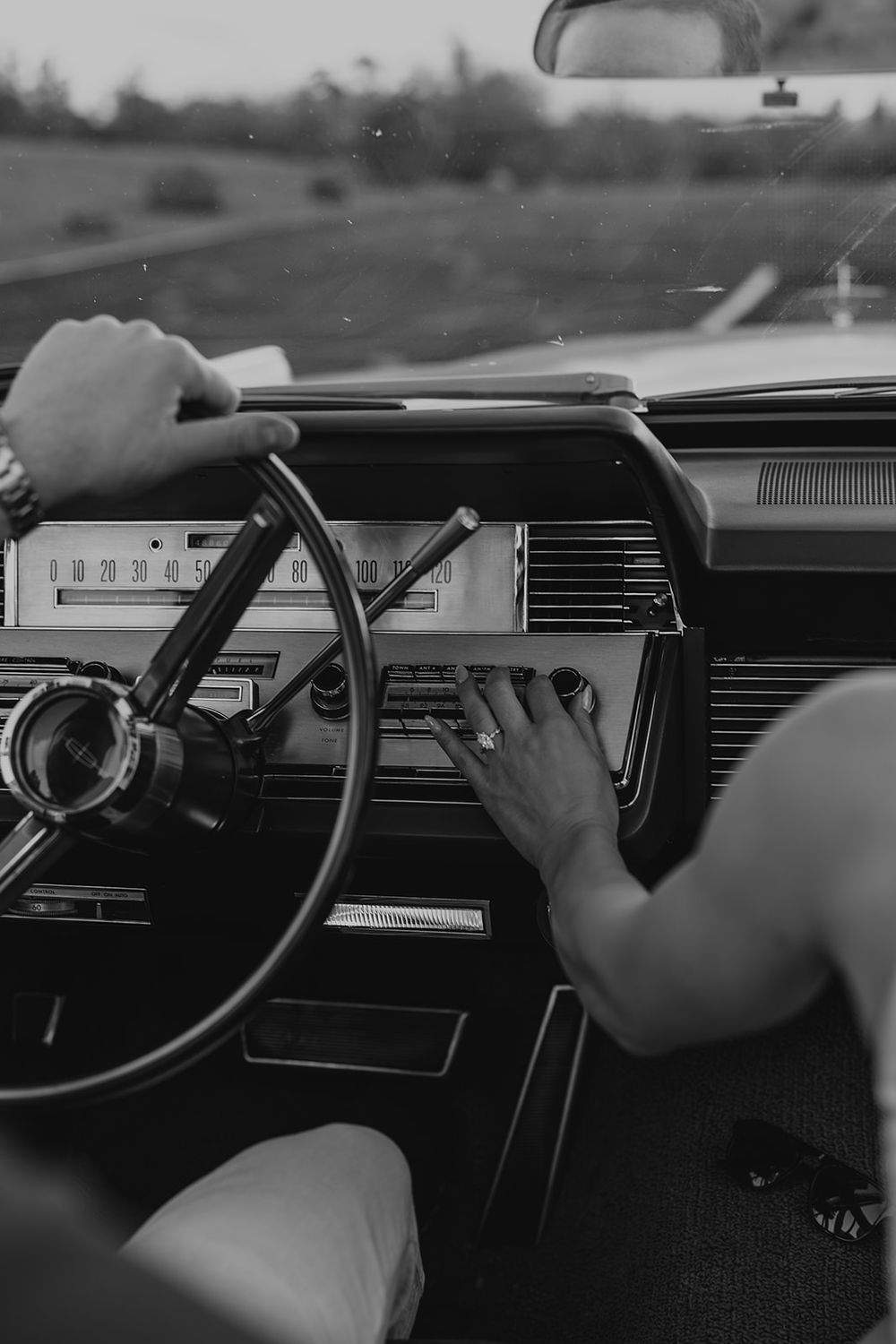 Black and white photo of a person adjusting a vintage car radio while driving, classic dashboard and steering wheel visible.