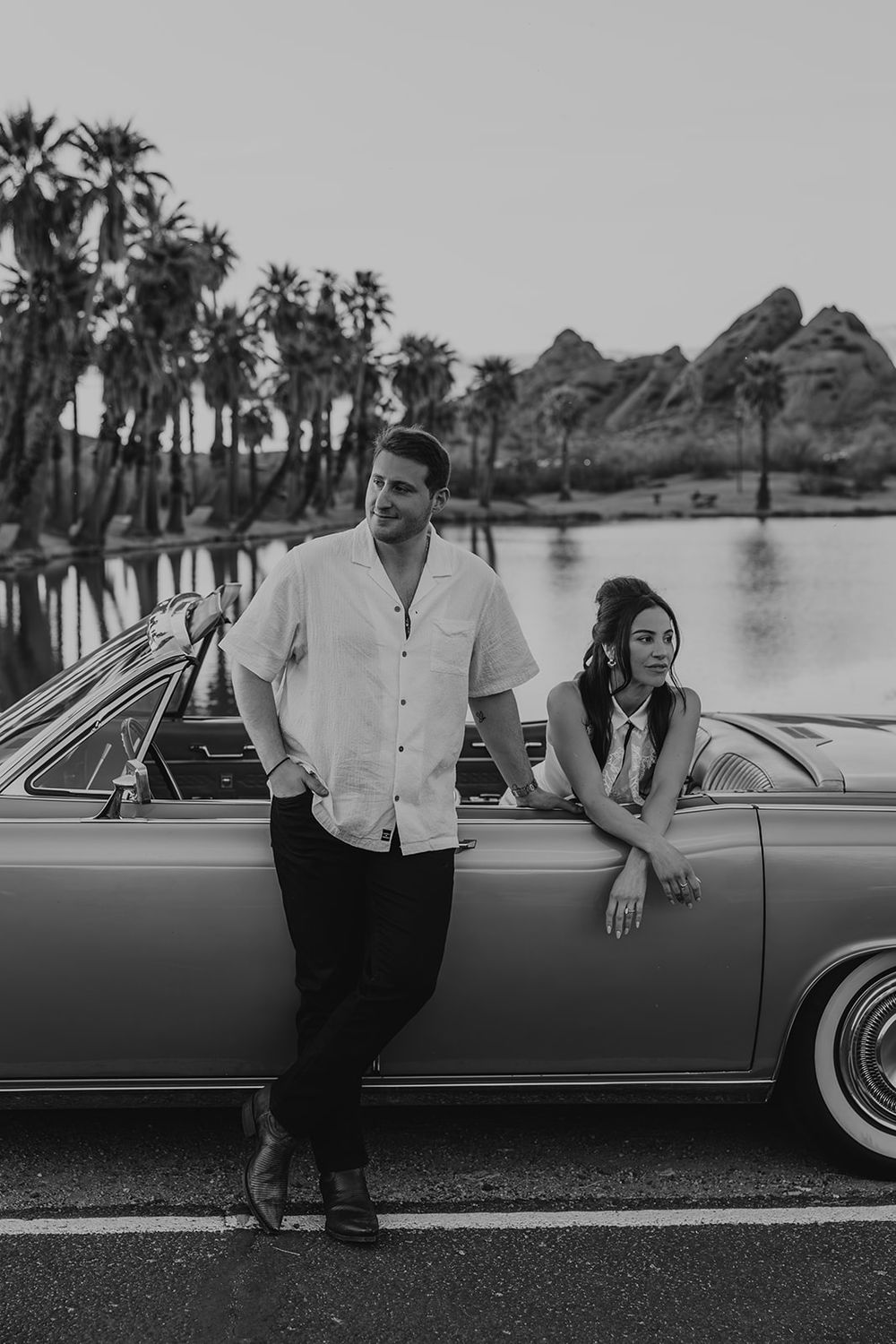 Black and white photo of a couple leaning against a vintage convertible car near a lake with palm trees and mountains.