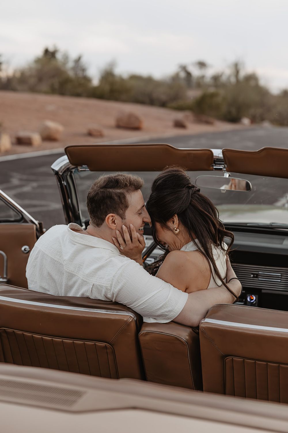 Couple sharing a romantic moment in a vintage convertible car with brown leather seats in a desert landscape.