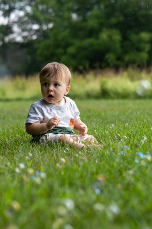 Curious baby sitting on lush green grass outdoors, wearing a white shirt, looking up with wonder on a sunny day.