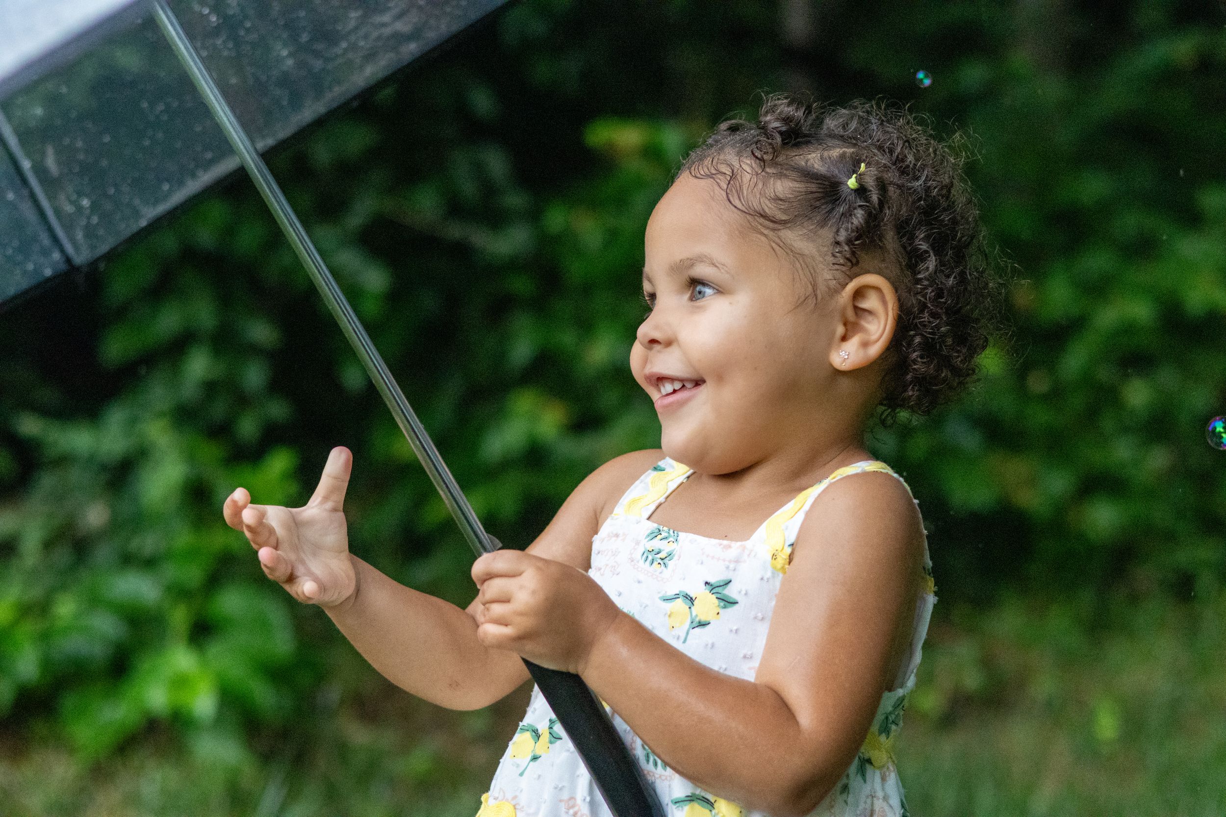 A joyful toddler girl holds an umbrella in the rain, wearing a floral dress, smiling outdoors.