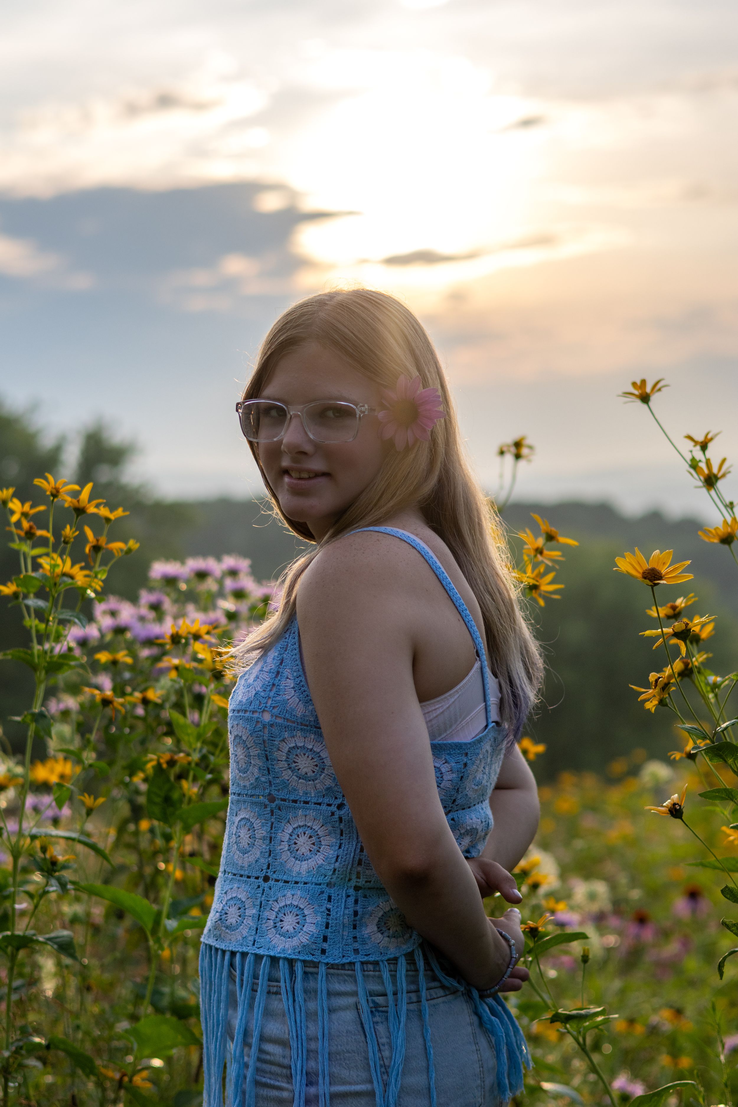 Young woman in blue dress and glasses smiling among wildflowers at golden sunset.