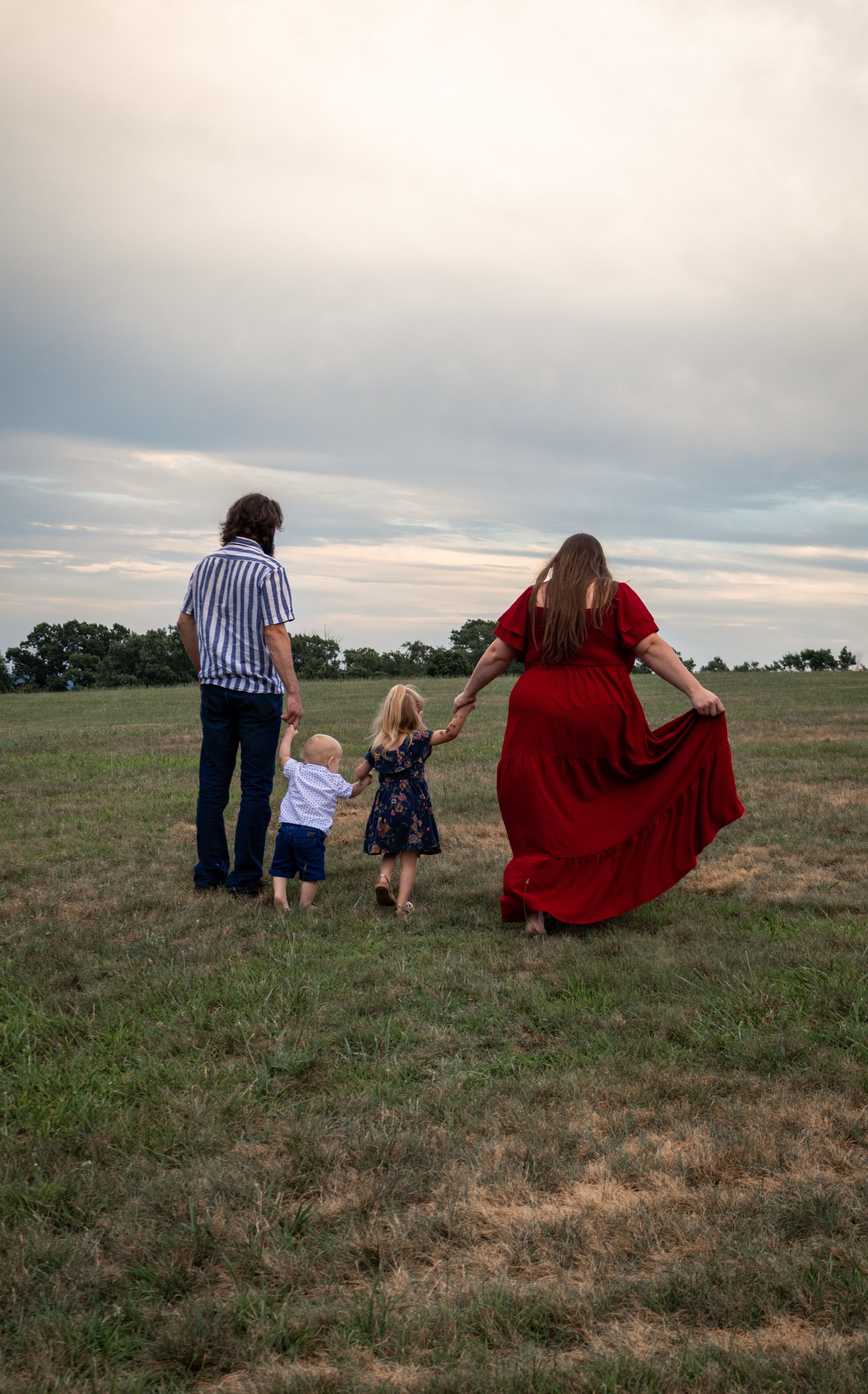 Family of four walking hand-in-hand across a green field at dusk, woman in flowing red dress, children between parents.