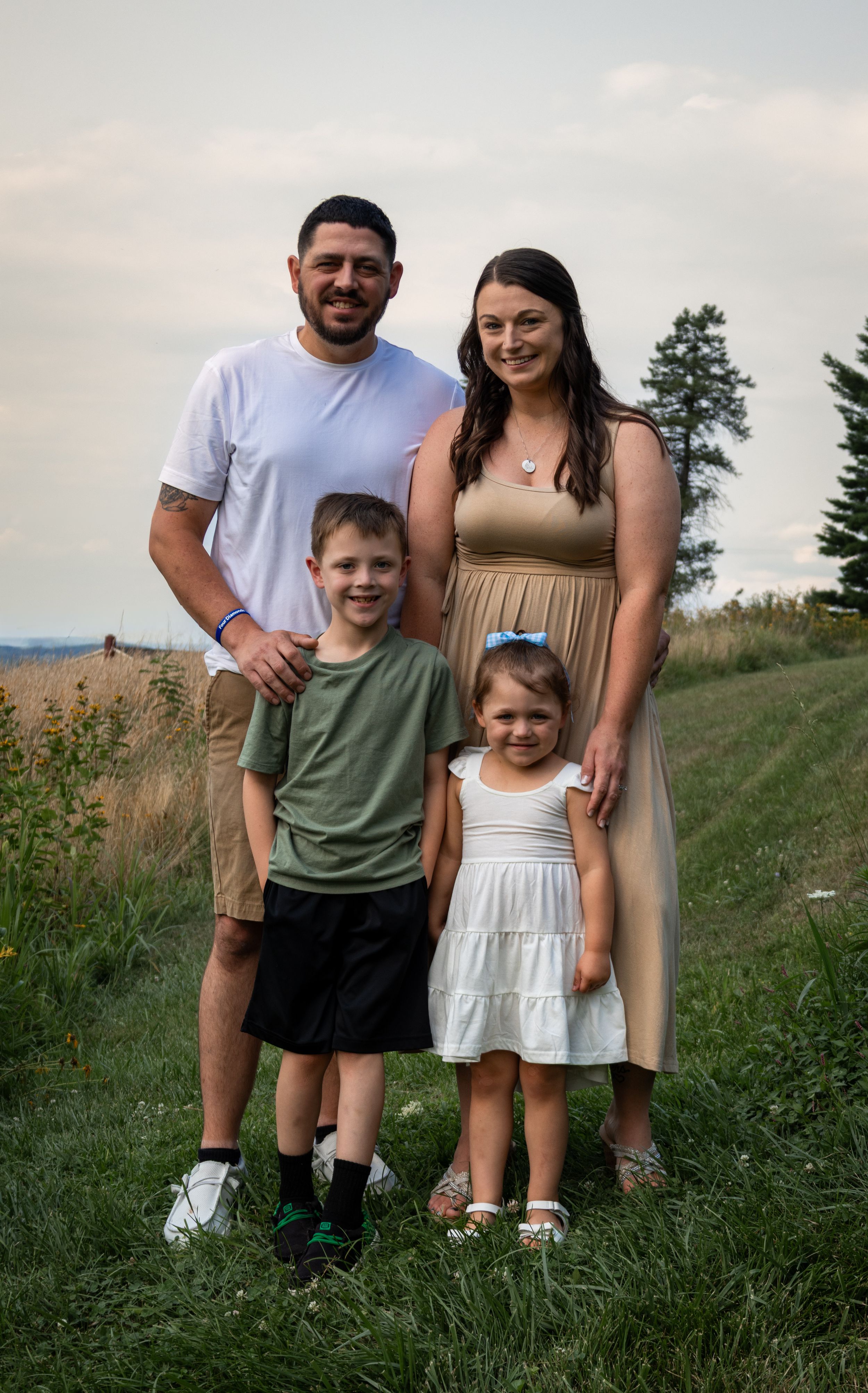 A happy family of four poses outdoors in a green field, with two adults and two young children smiling for a portrait.