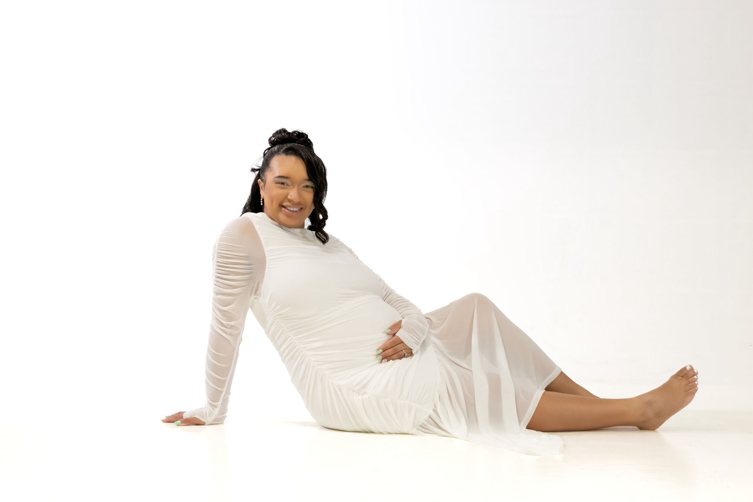 Pregnant woman in white sheer dress sitting gracefully on floor, smiling, studio photo on white background.