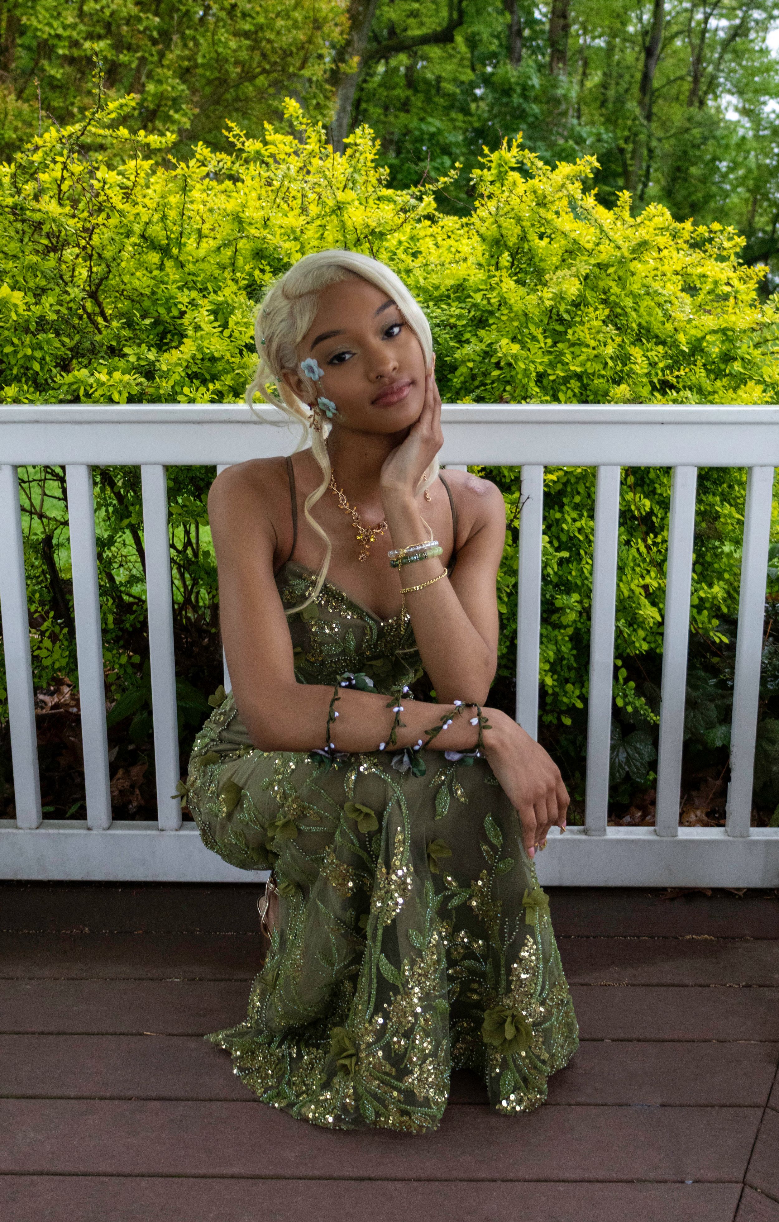 Young woman in green sequin gown sits on wooden deck surrounded by lush greenery, wearing gold jewelry and blonde updo.