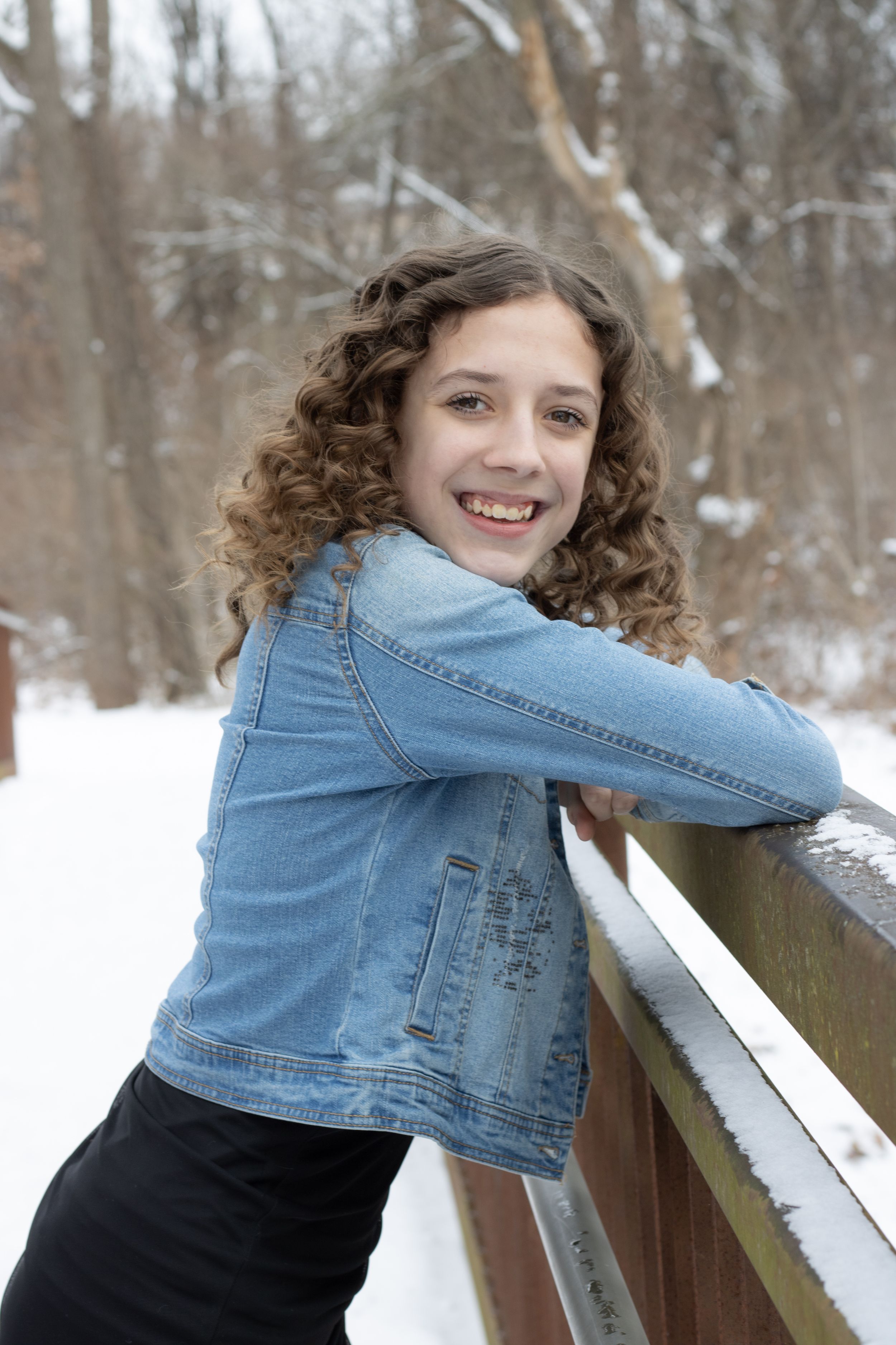 Smiling teenage girl with curly hair in denim jacket leaning on snowy wooden railing in winter forest.