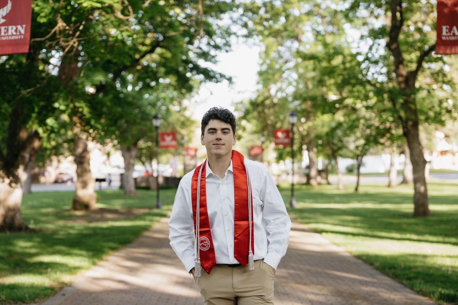 College graduate in white shirt and red sash stands on tree-lined campus walkway with red university banners.