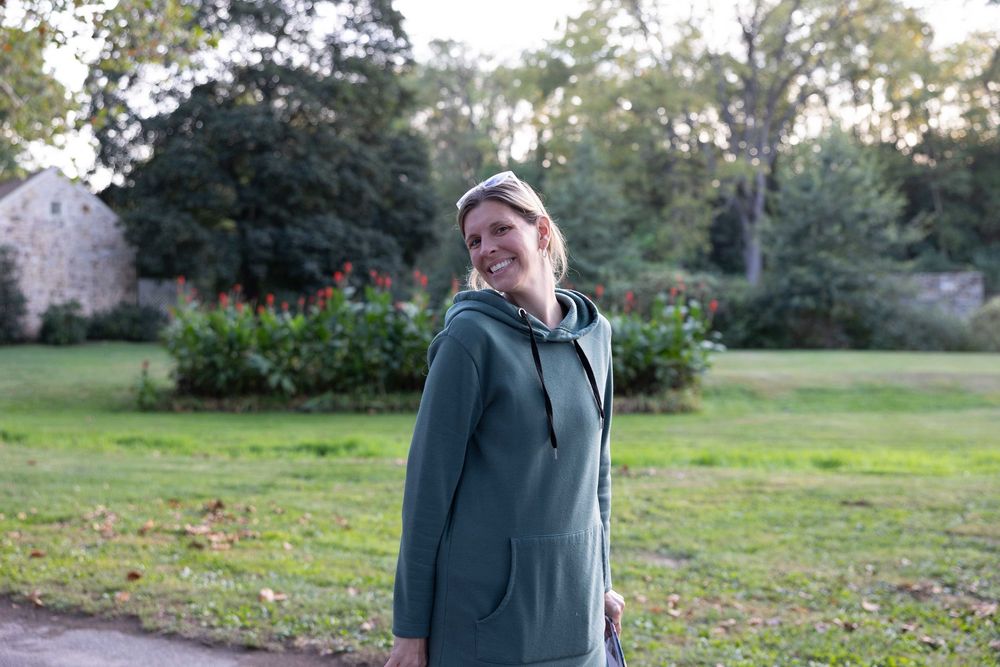Someone wearing a green hooded coat smiles while standing in a garden setting on an autumn day.