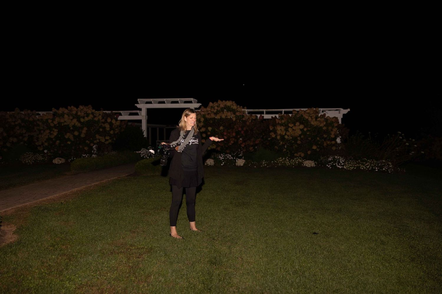 A person stands on a dark lawn at night wearing a black outfit and holding an object.
