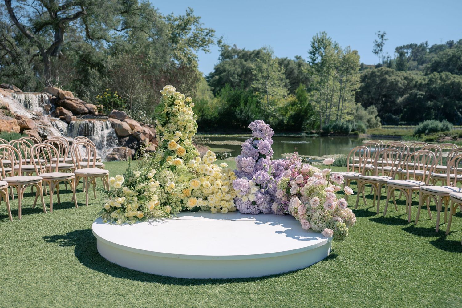 Elegant outdoor wedding ceremony setup with white chairs, floral arrangements, and a curved aisle on a manicured lawn.