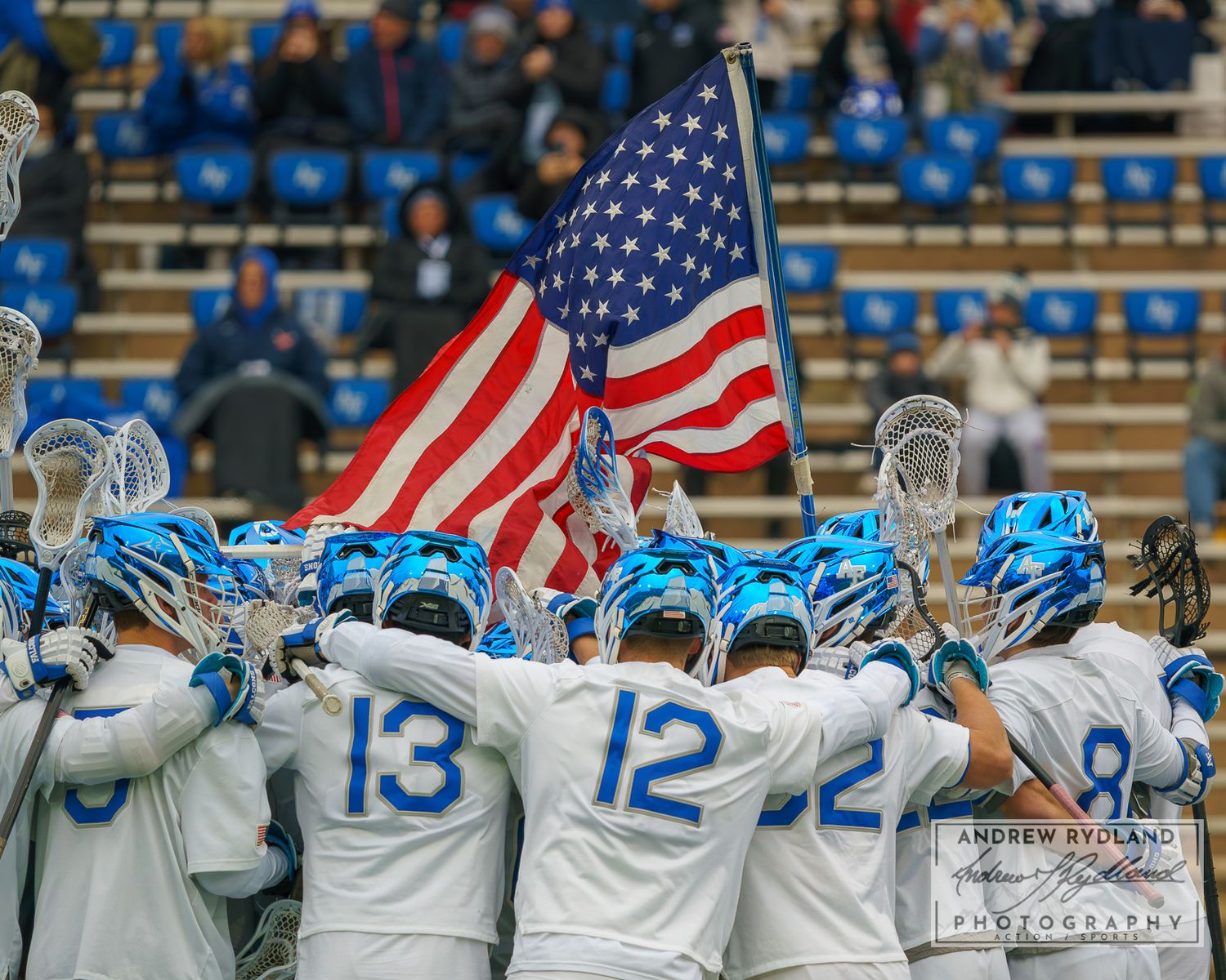 College lacrosse team huddles together with American flag displayed during pre-game ceremony.