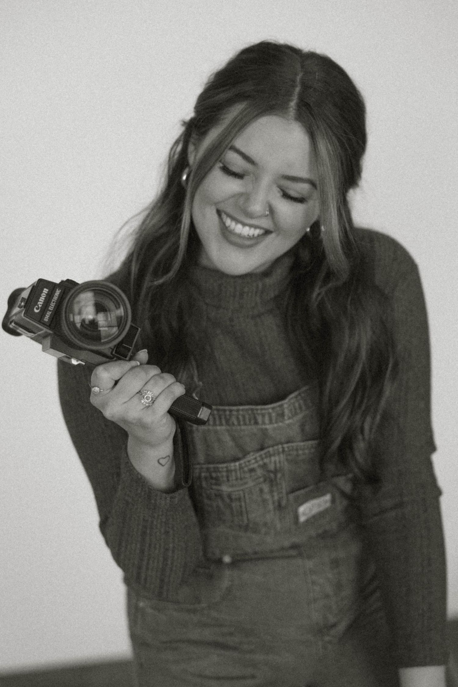 Smiling woman in overalls holding Canon camera in black and white portrait photography shot.