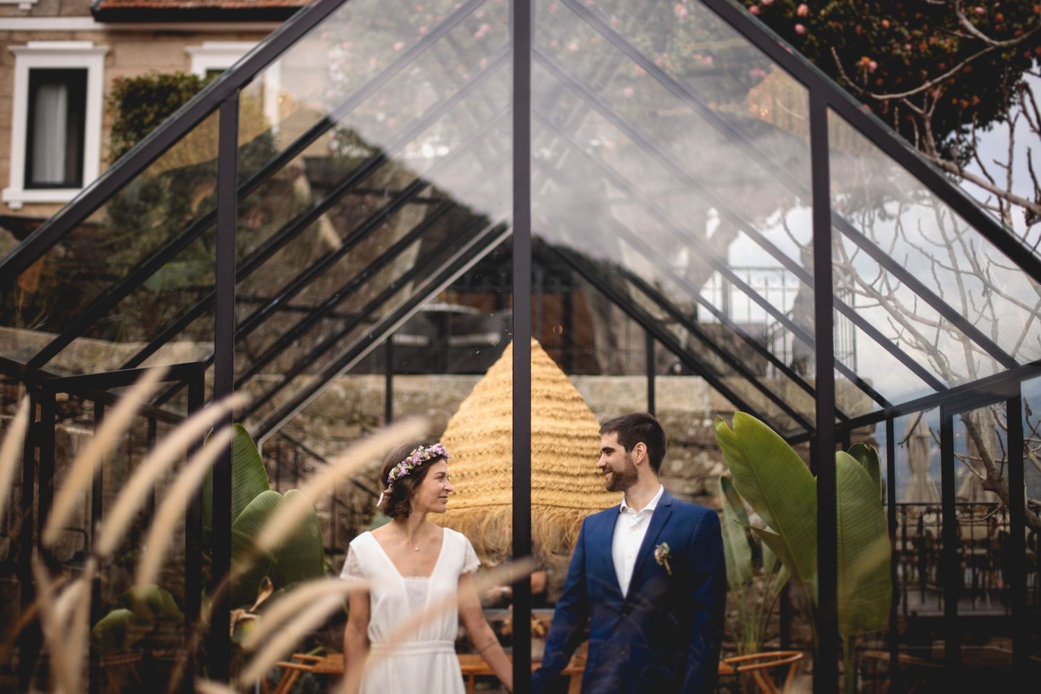 Bride and groom share an intimate moment under a greenhouse glass ceiling with sunlight streaming through.