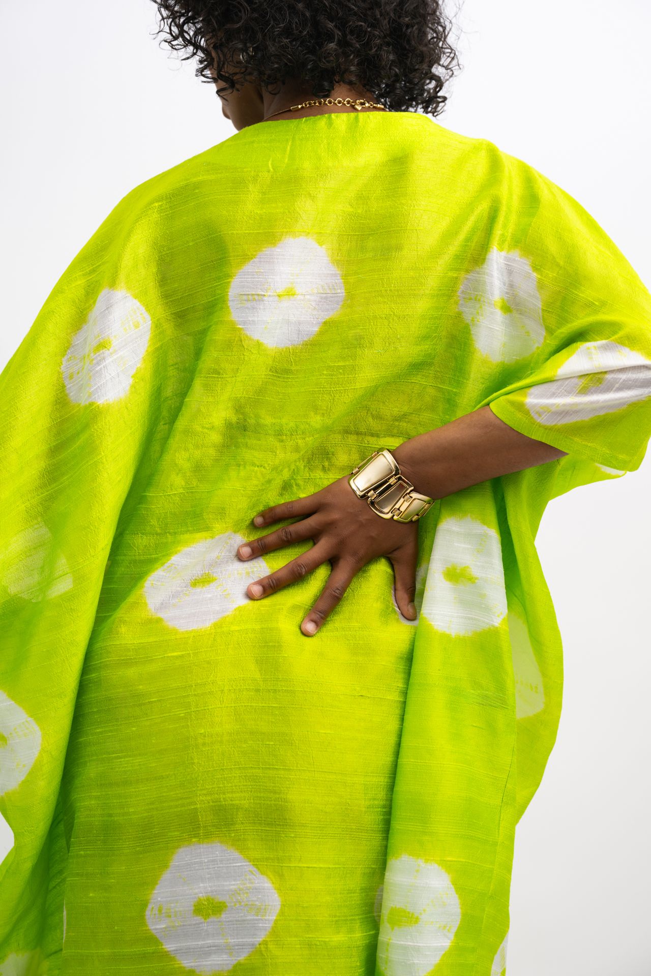 Person wearing a neon green kaftan with white polka dots, gold bracelet and necklace, back view.