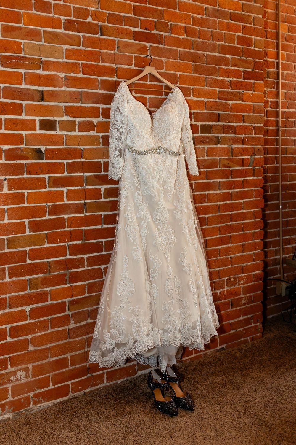 An elegant white lace wedding dress with 3/4 sleeves hangs against a rustic red brick wall
