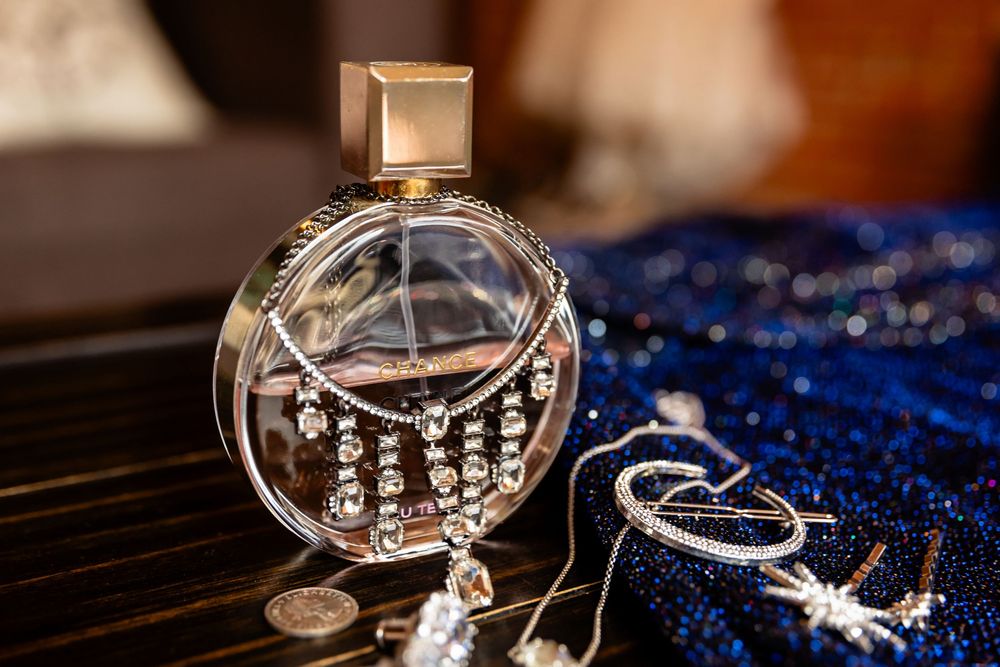 Elegant crystal perfume bottle with rhinestone details beside blue sequin fabric and silver jewelry on dark surface.