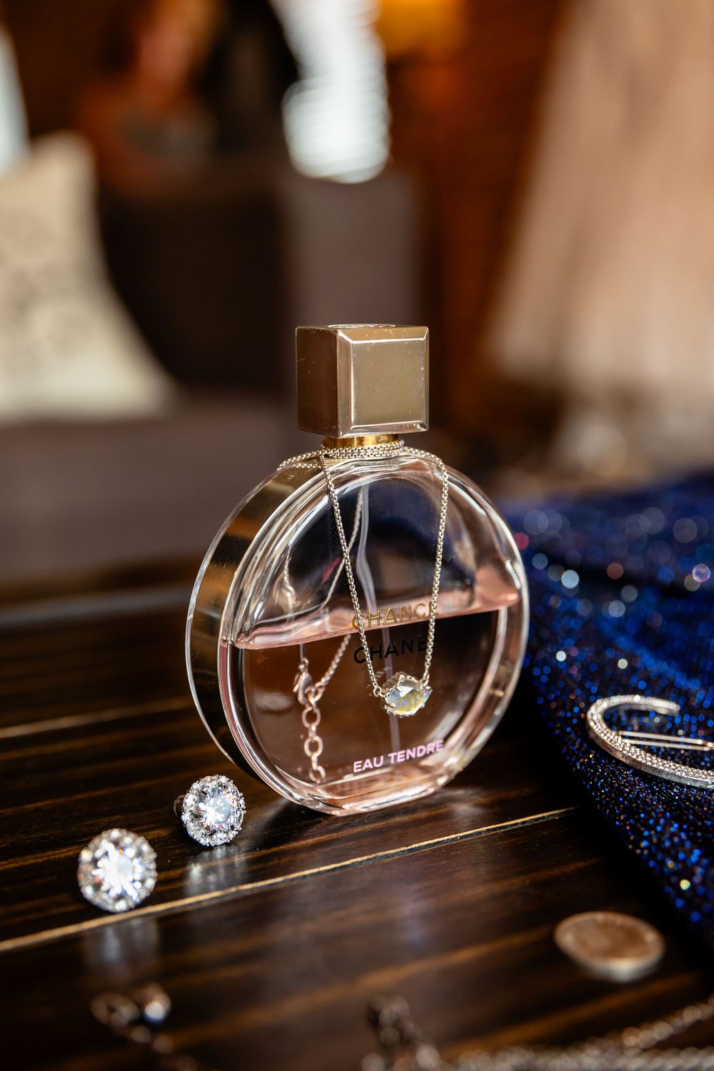 Round glass perfume bottle with gold cap, diamond earrings, and delicate necklaces on a wooden surface.