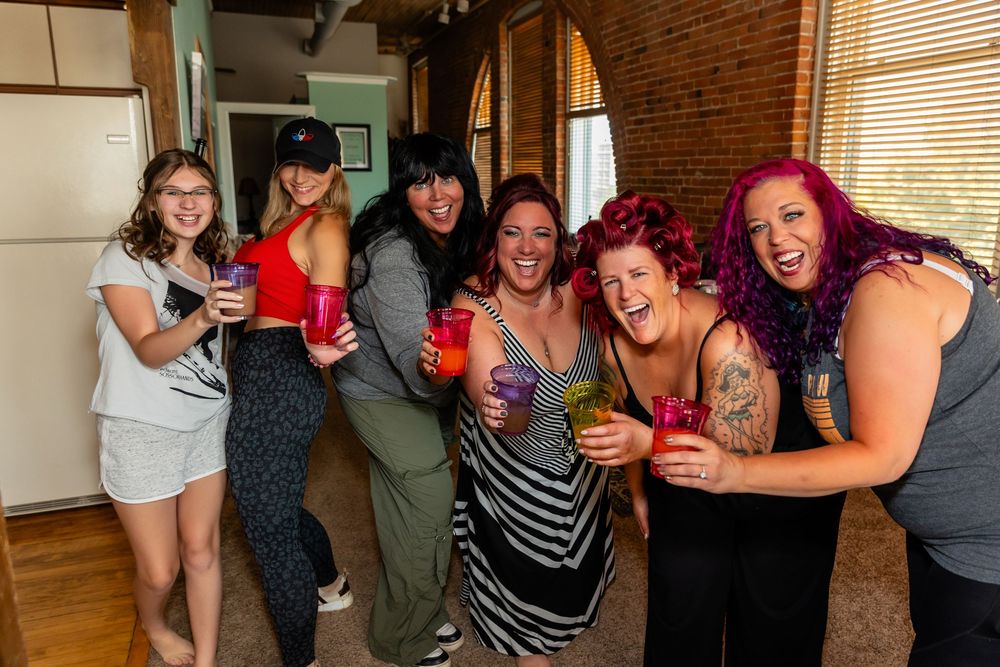 Six women laughing and posing together holding colorful drinks at a lively indoor gathering.