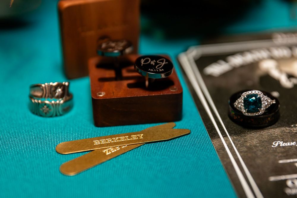 Wedding details featuring monogrammed cufflinks, silver rings, gold collar stays, and a teal gemstone ring on a turquoise surface.