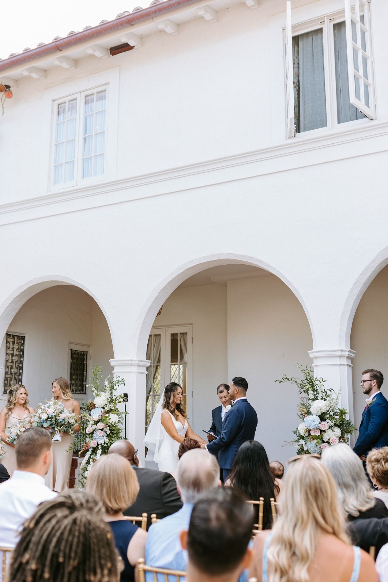 Outdoor wedding ceremony in a white Mediterranean courtyard with floral arrangements and guests seated in gold chairs at Darlington House.
