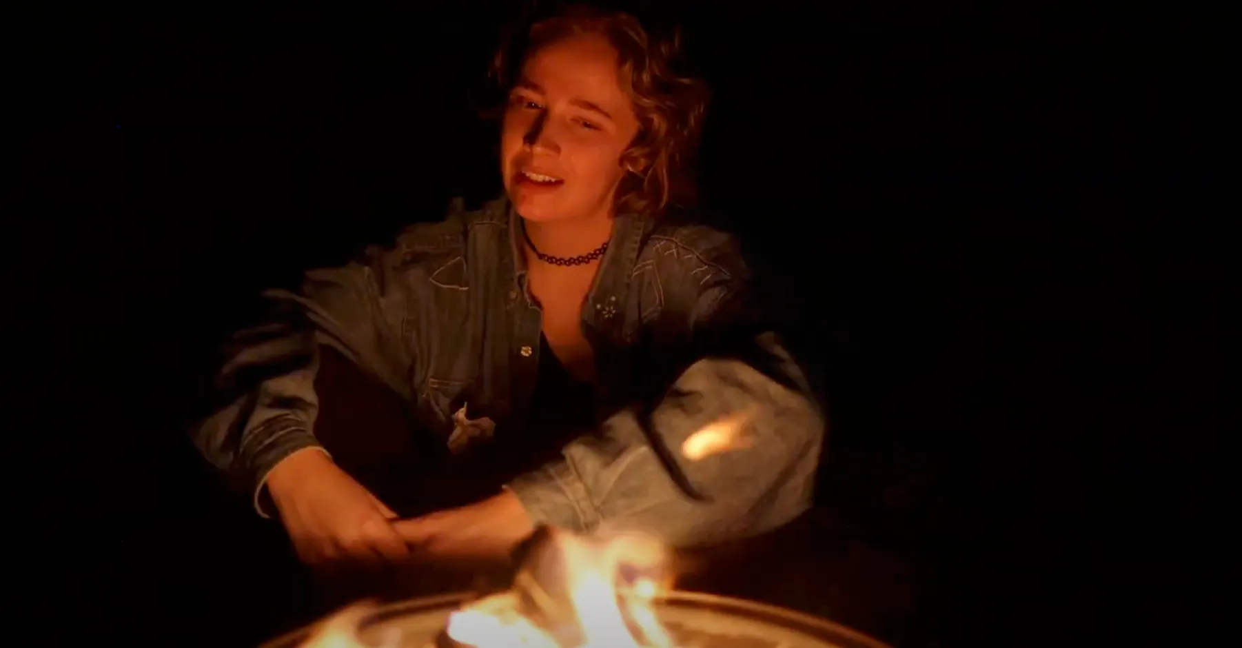 Person smiling warmly while tending to a glowing fire bowl in a dark outdoor setting.