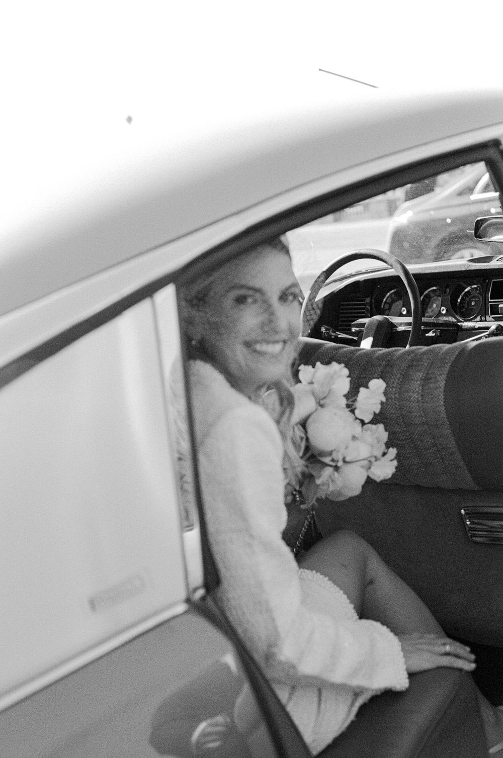 Smiling bride holding a bouquet of flowers sitting in a vintage car on her wedding day, black and white photo.