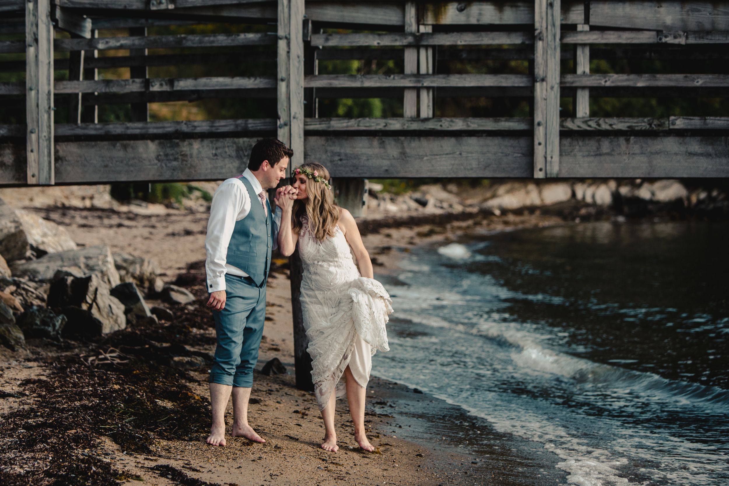 Barefoot bride and groom share a romantic moment on a rocky shoreline near a weathered wooden structure.