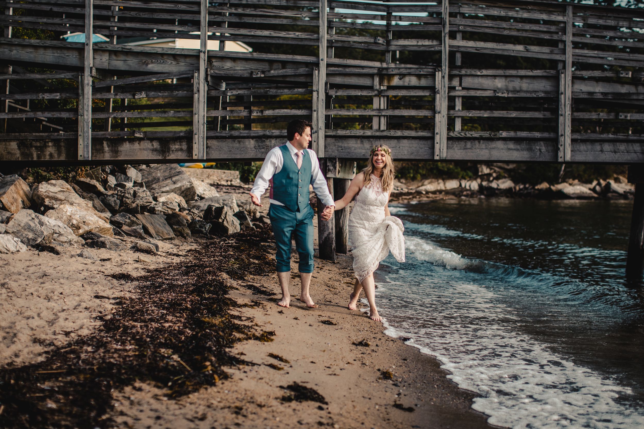 Bride and groom walk barefoot along a sandy beach shoreline near a wooden bridge on their wedding day.