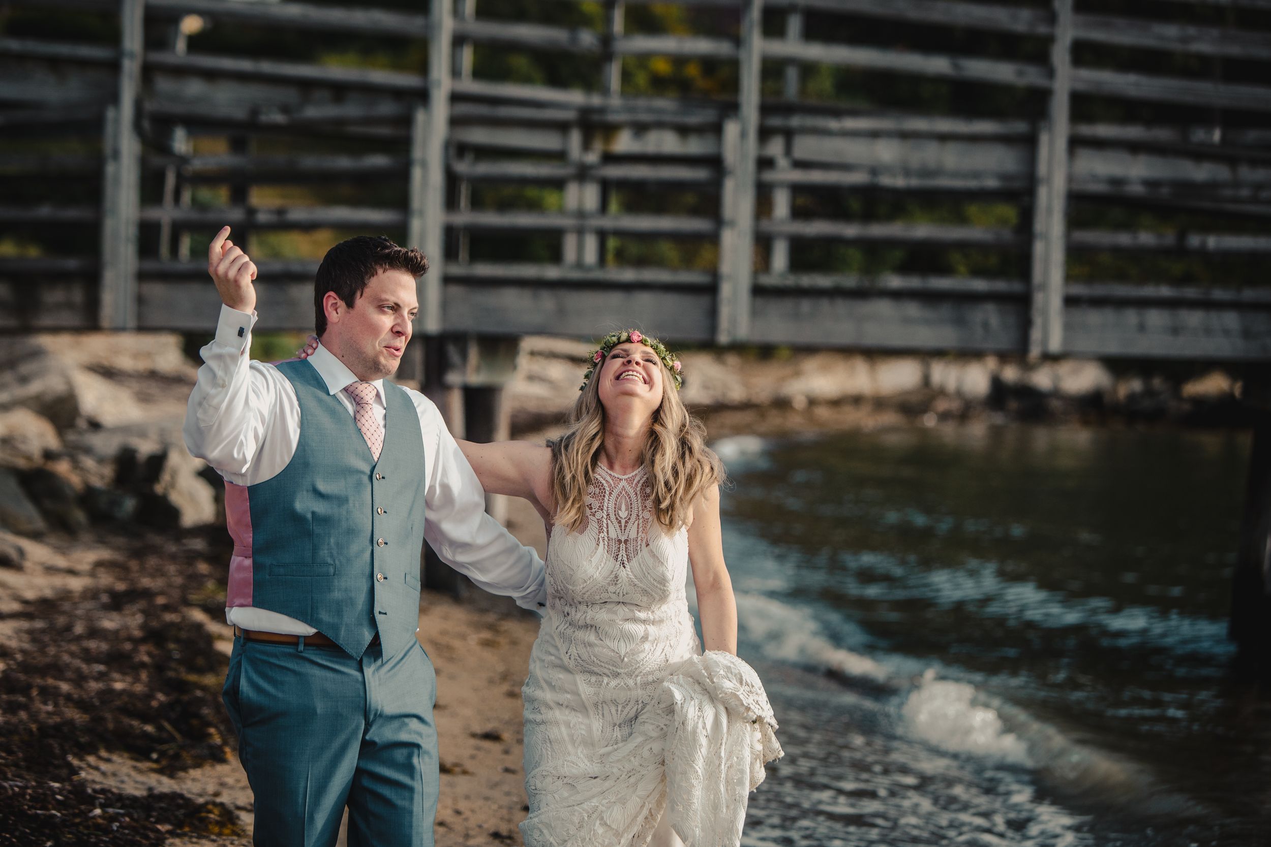 Bride and groom walking by waterfront in wedding attire, groom in blue suit tossing item while bride laughs joyfully.