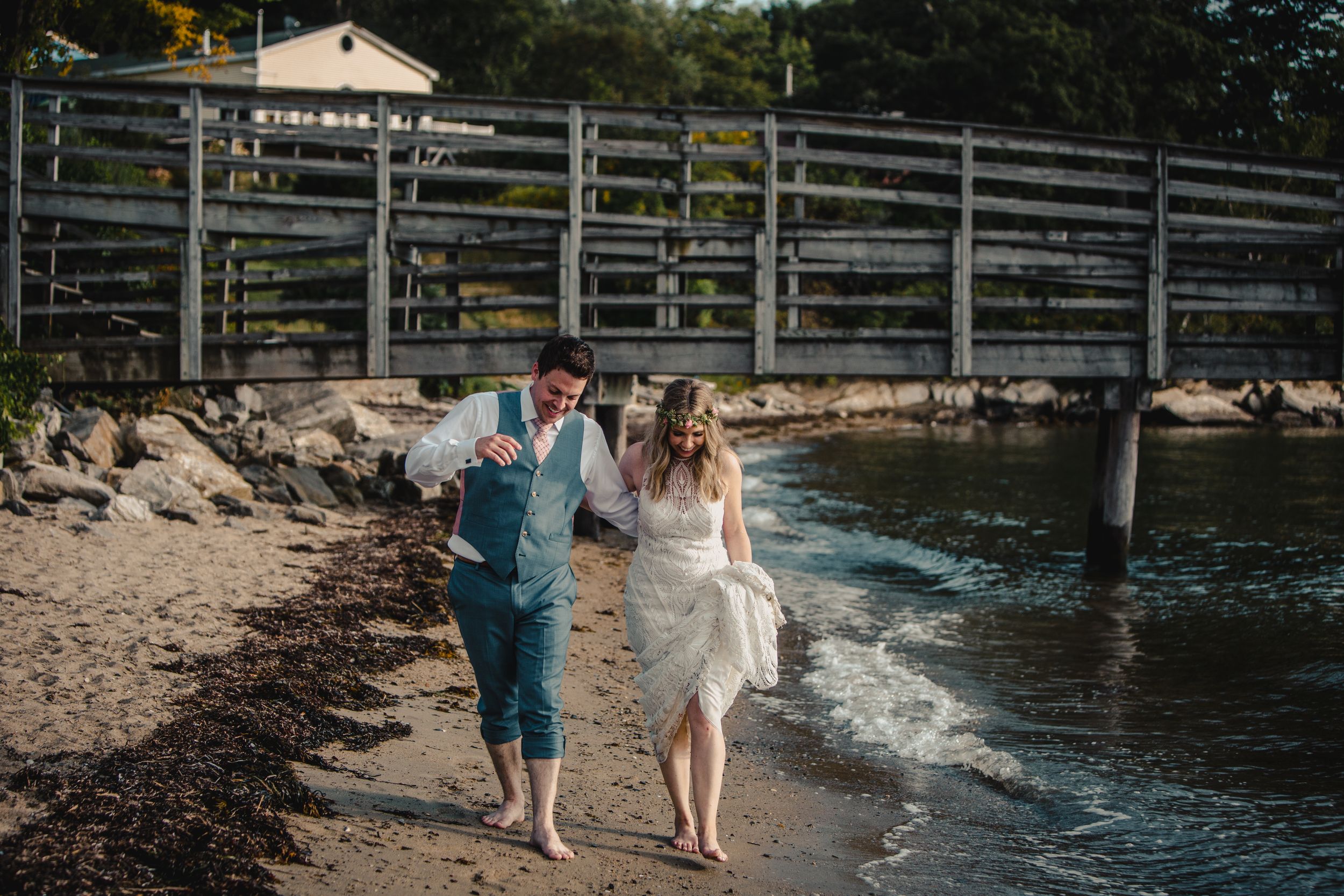Bride and groom walking barefoot along a sandy beach shore near a wooden bridge on their wedding day.