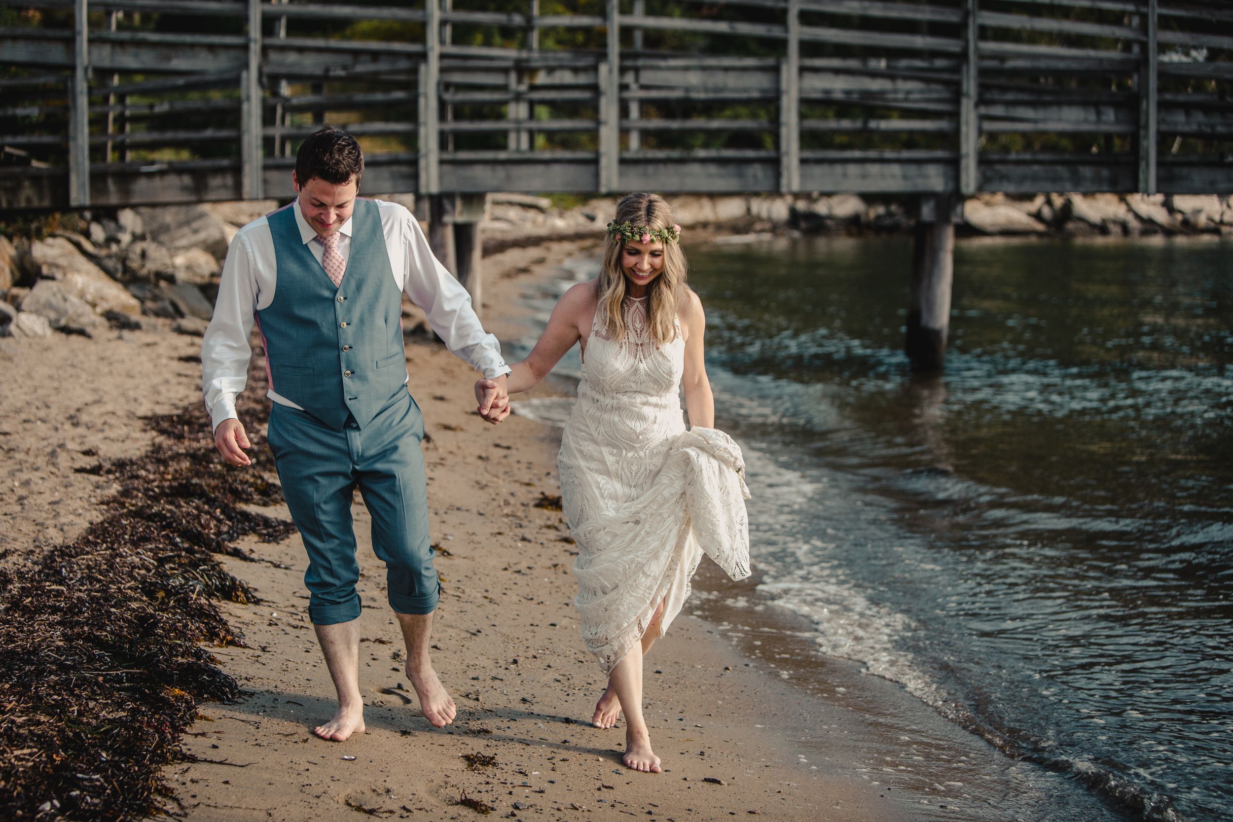 Barefoot bride and groom walk hand in hand along sandy beach shoreline on their wedding day.